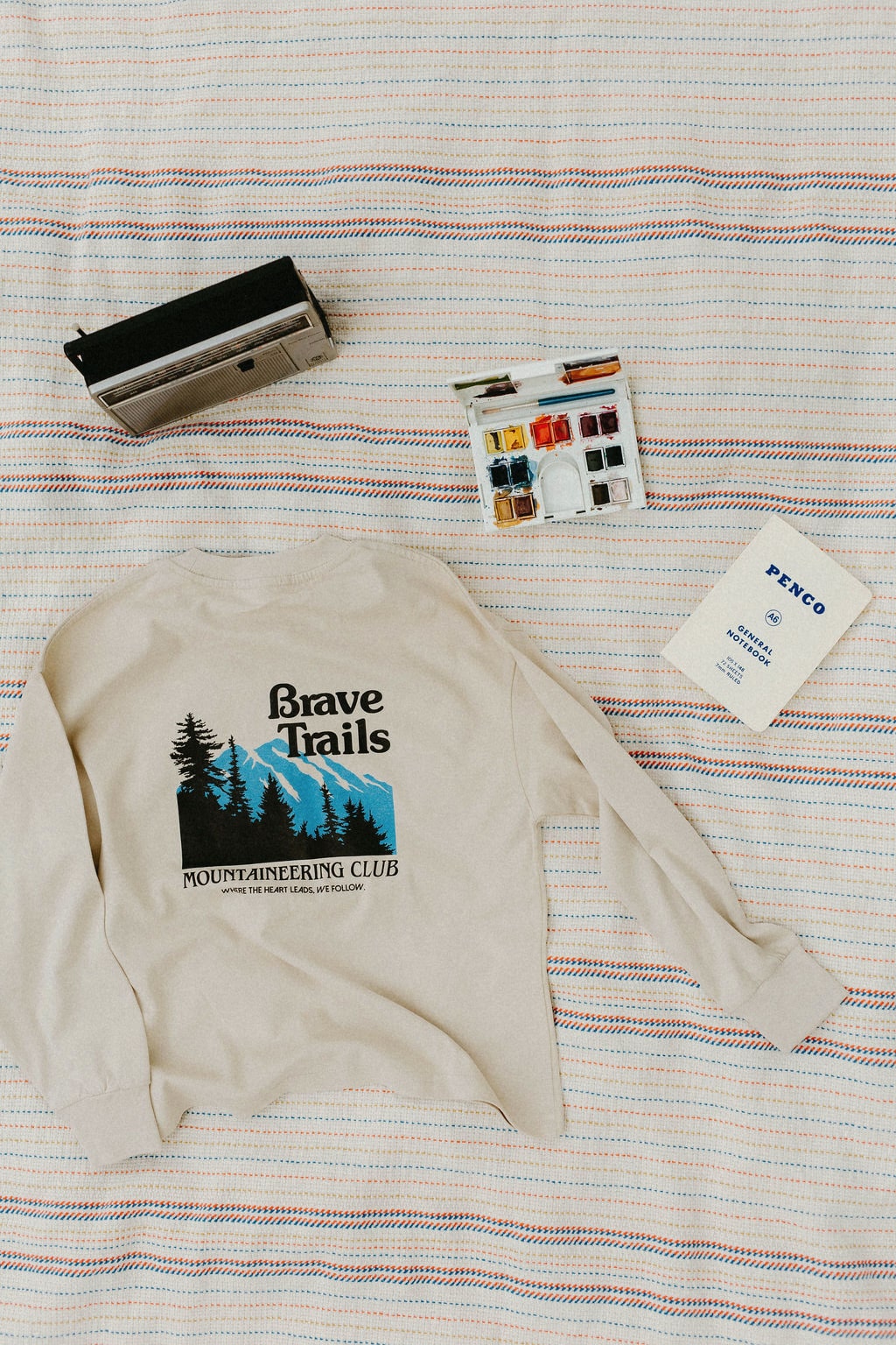 Back of a beige long sleeve t-shirt with 'Brave Trails Mountaineering Club' design on a striped blanket background