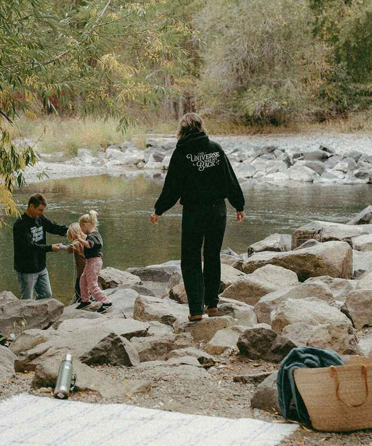 Two adults and a child walking along a rocky riverbank with trees in the background.