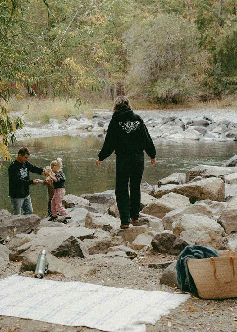 Two adults and a child walking along a rocky riverbank with trees in the background.