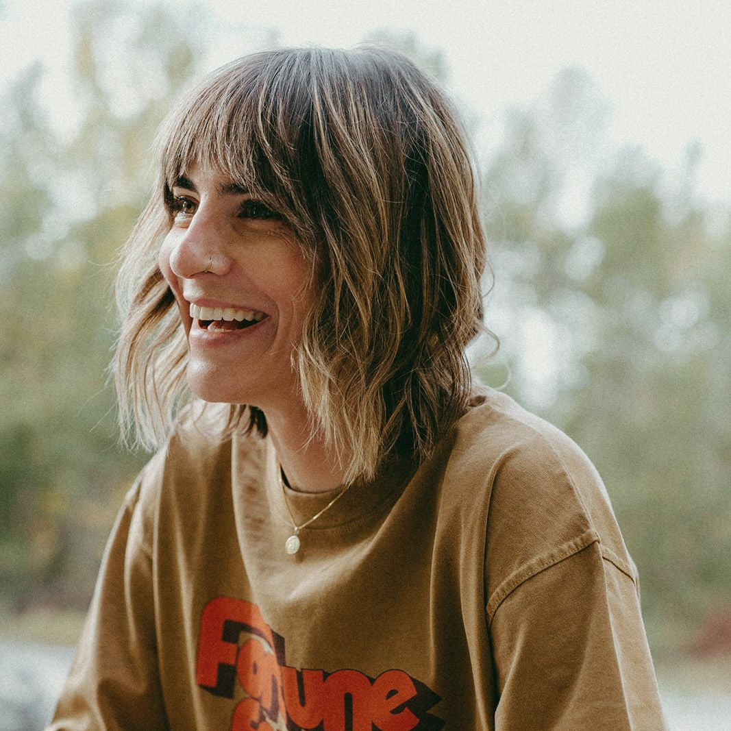 Woman wearing a brown t-shirt with red text, sitting outdoors. T-shirt says "fortune favors the bold"