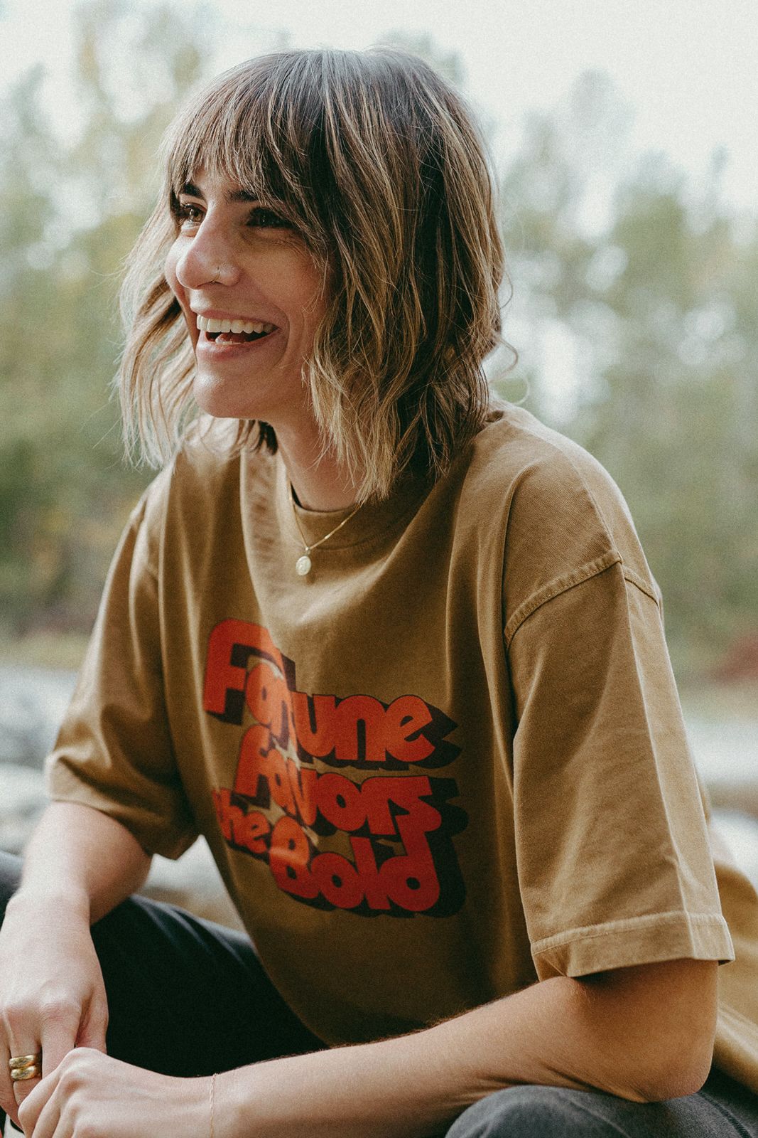 Woman wearing a brown t-shirt with red text, “fortune favors the bold” sitting outdoors.
