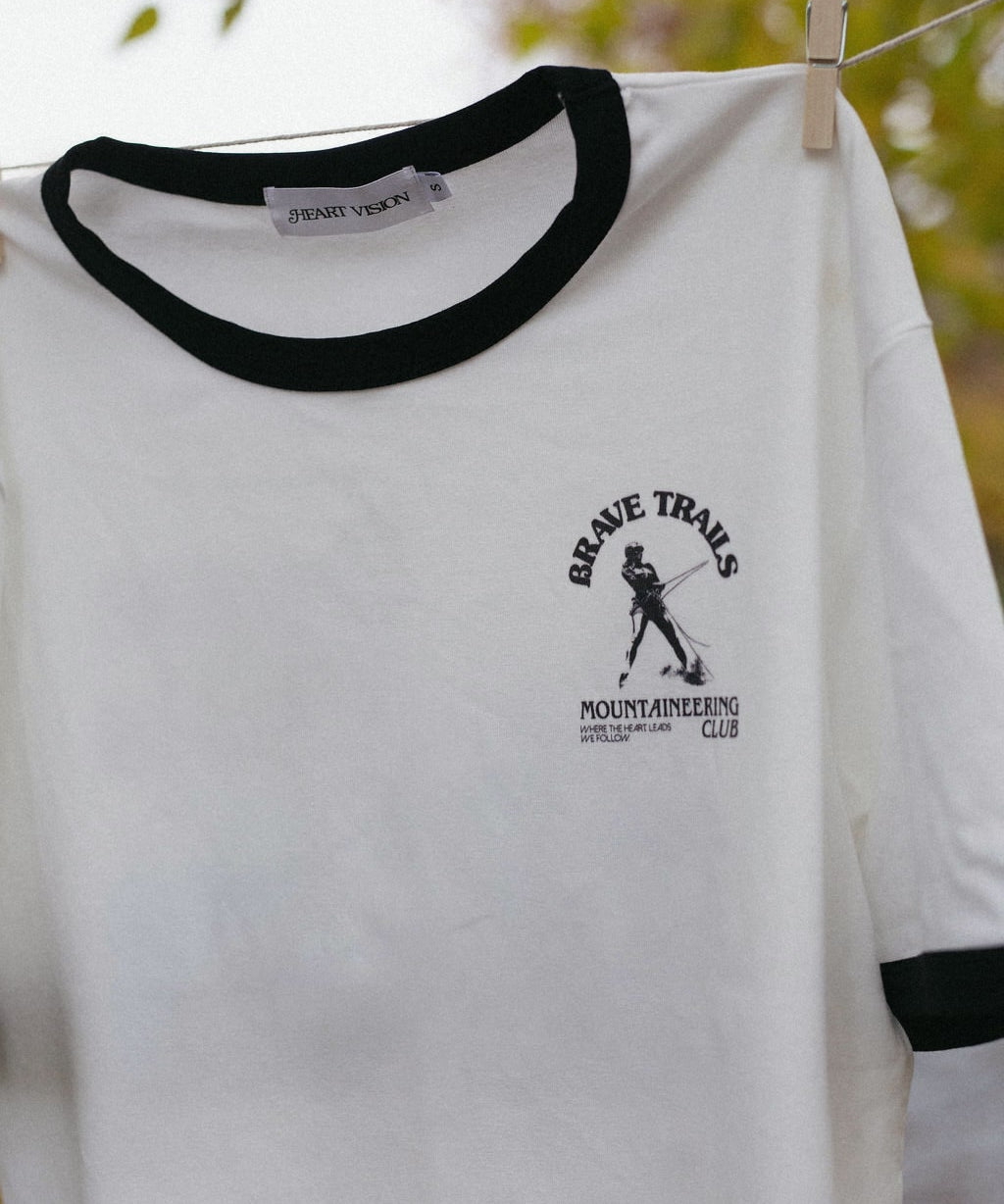 White t-shirt with black trim hanging on a clothesline, featuring a brave trails mountaineering logo with a blurred natural background