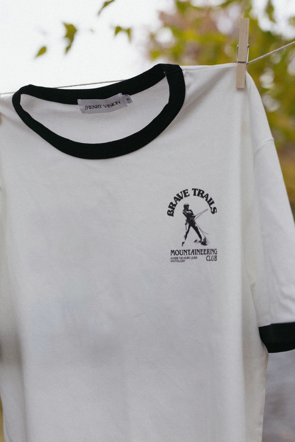 White t-shirt with black trim hanging on a clothesline, featuring a brave trails mountaineering logo with a blurred natural background