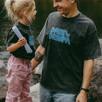Man and child standing by a body of water, both wearing black t-shirts with 'Dare to Dream' text.