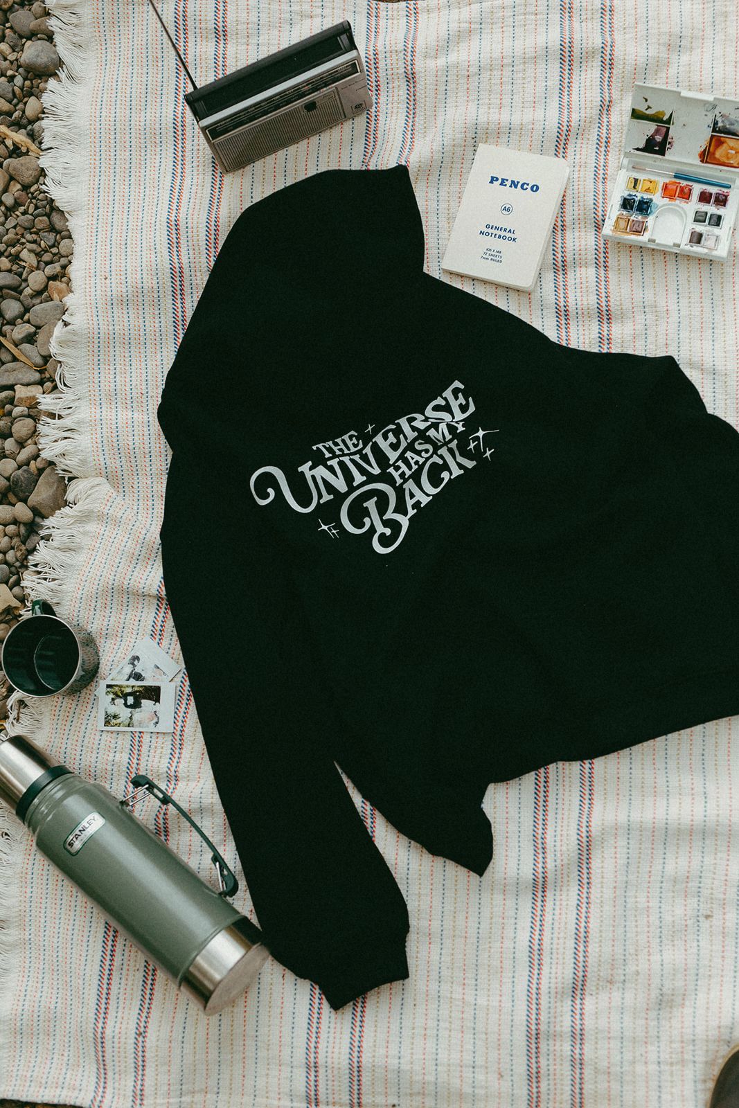 Black sweatshirt with text on a blanket with camping gear around and art supplies