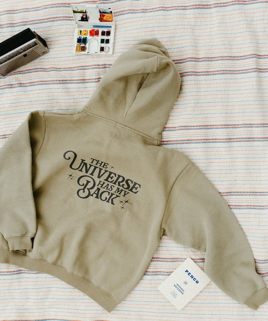 Khaki hoodie with 'The Universe Has My Back' text on a striped fabric background