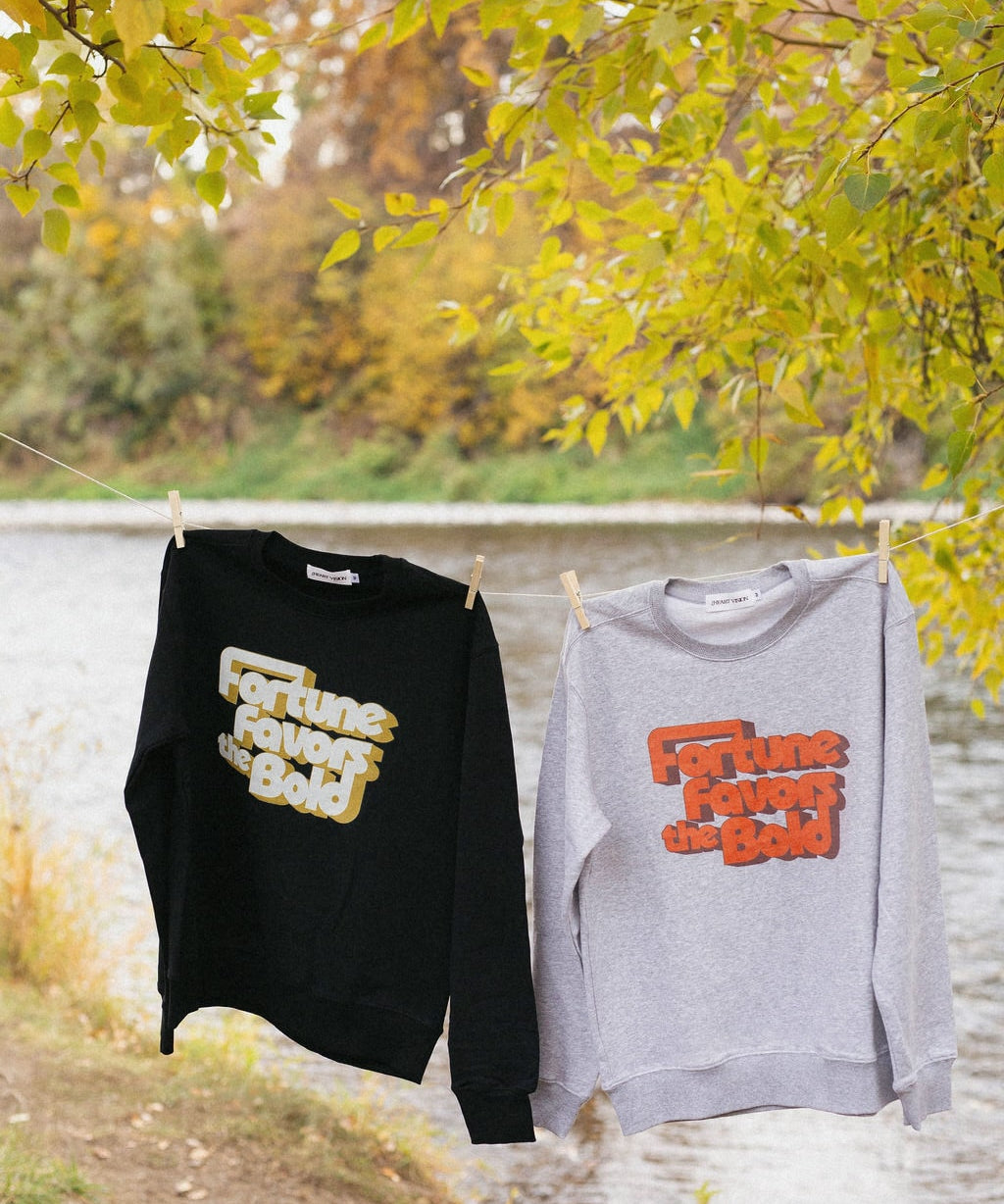 Two sweatshirts with text "fortune favors the bold" hanging on a clothesline by a body of water with autumn foliage.