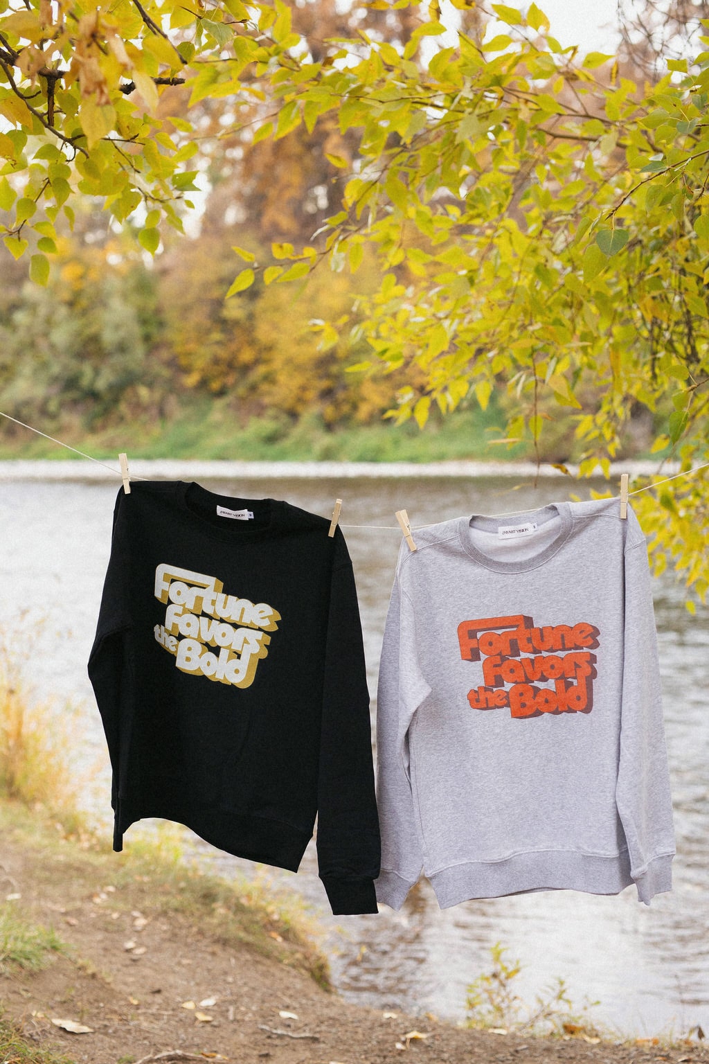 Two sweatshirts with text "fortune favors the bold" hanging on a clothesline by a body of water with autumn foliage.