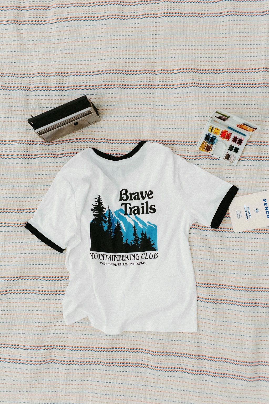 Back of white t-shirt with mountain graphic and Brave Trails Mountaineering text on a striped blanket background