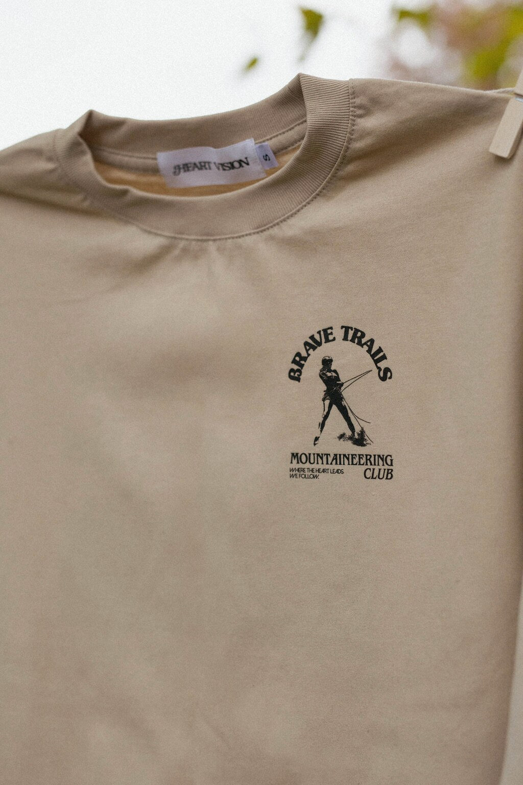 Close up view of "Brave Trails Mountaineering" logo on a beige long sleeve t-shirt on a blurred natural background