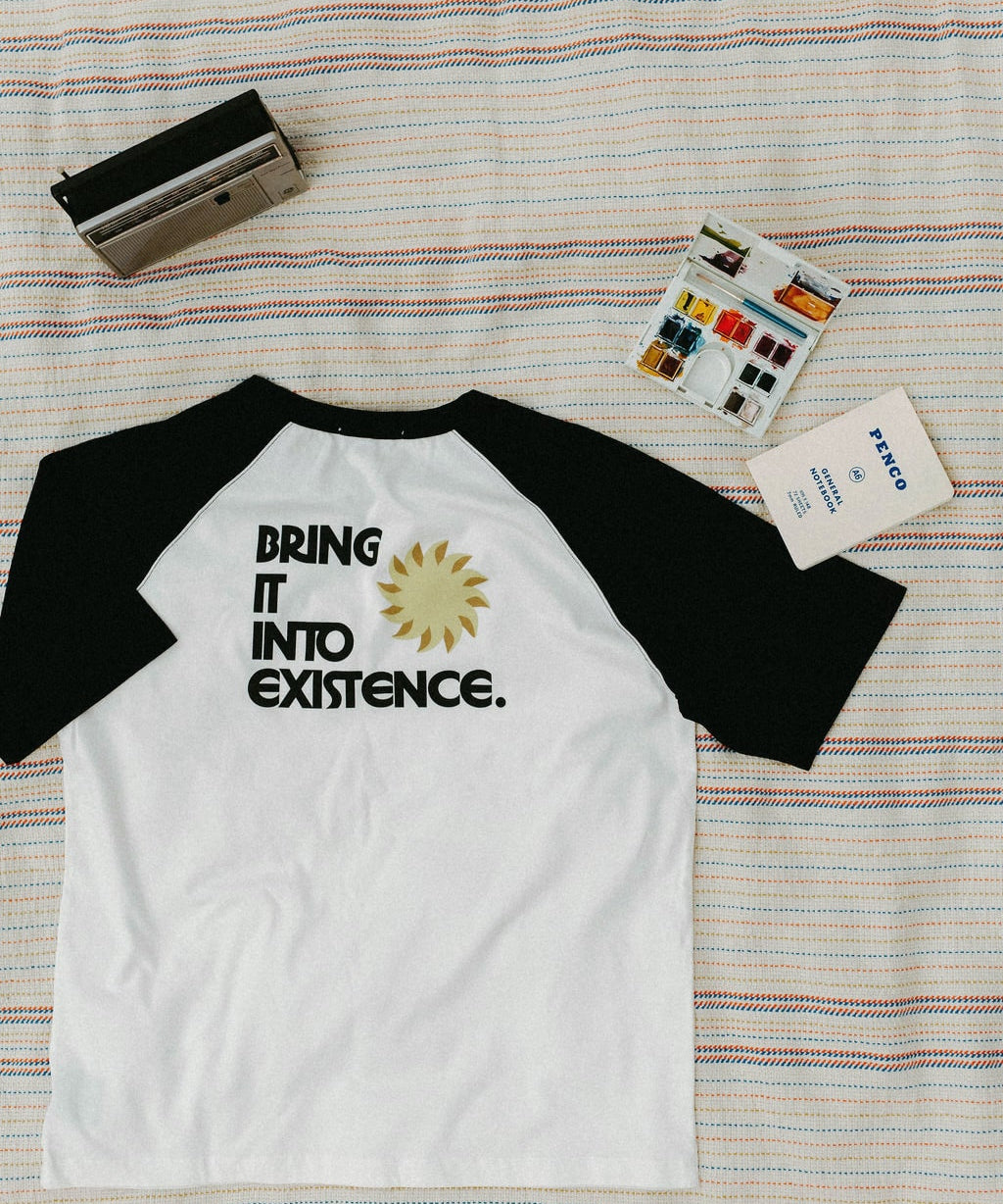 Back of white and black baseball shirt with 'Bring it into existence' text on a striped blanket background