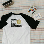 Back of white and black baseball shirt with 'Bring it into existence' text on a striped blanket background
