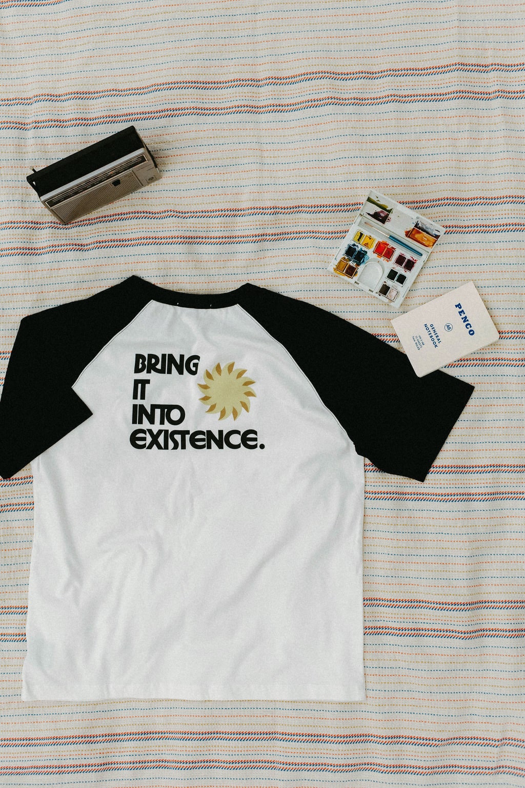 Back of white and black baseball shirt with 'Bring it into existence' text on a striped blanket background