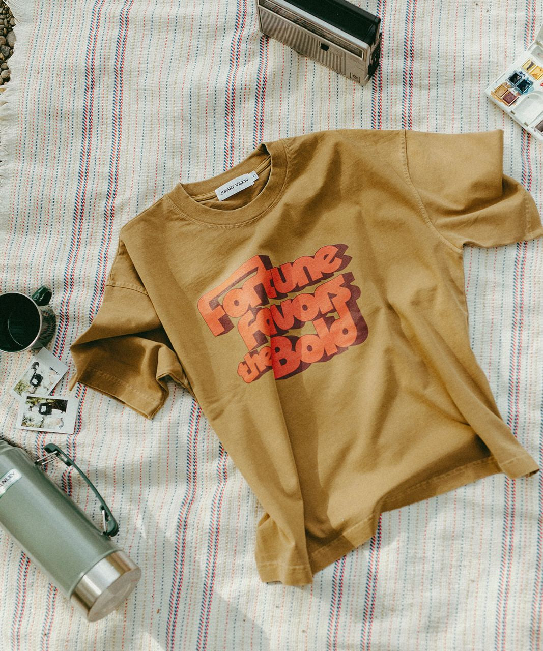 Brown t-shirt with red text on a striped blanket with a thermos and camera.