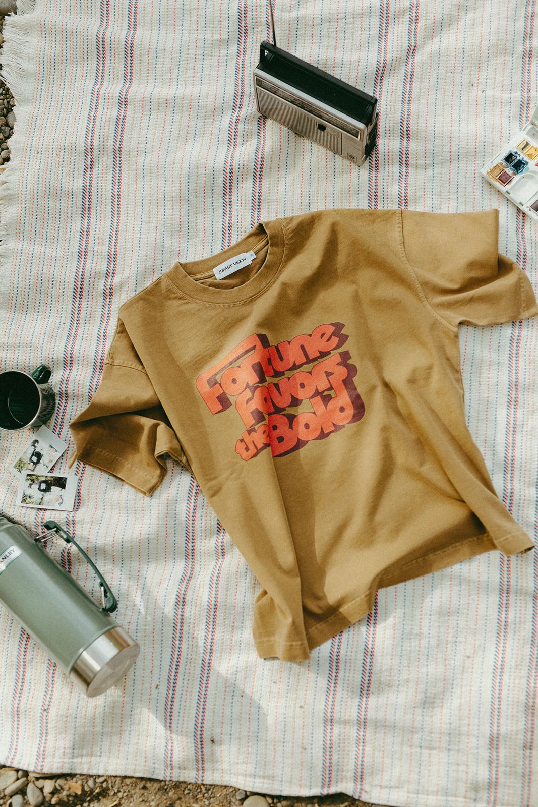 Brown t-shirt with red text on a striped blanket with a thermos and camera.