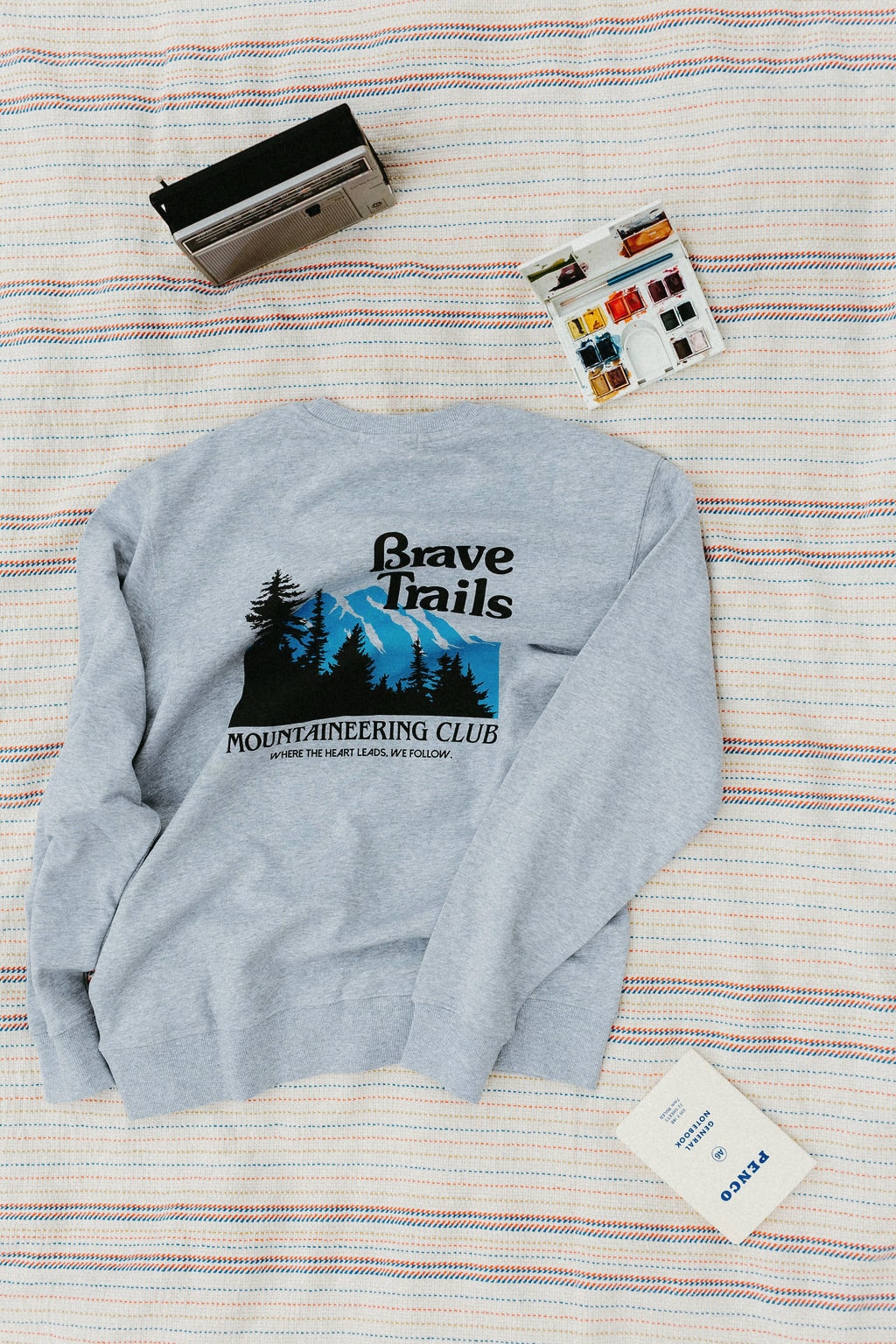 Gray sweatshirt with 'Brave Trails Mountaineering Club' design on a striped blanket background