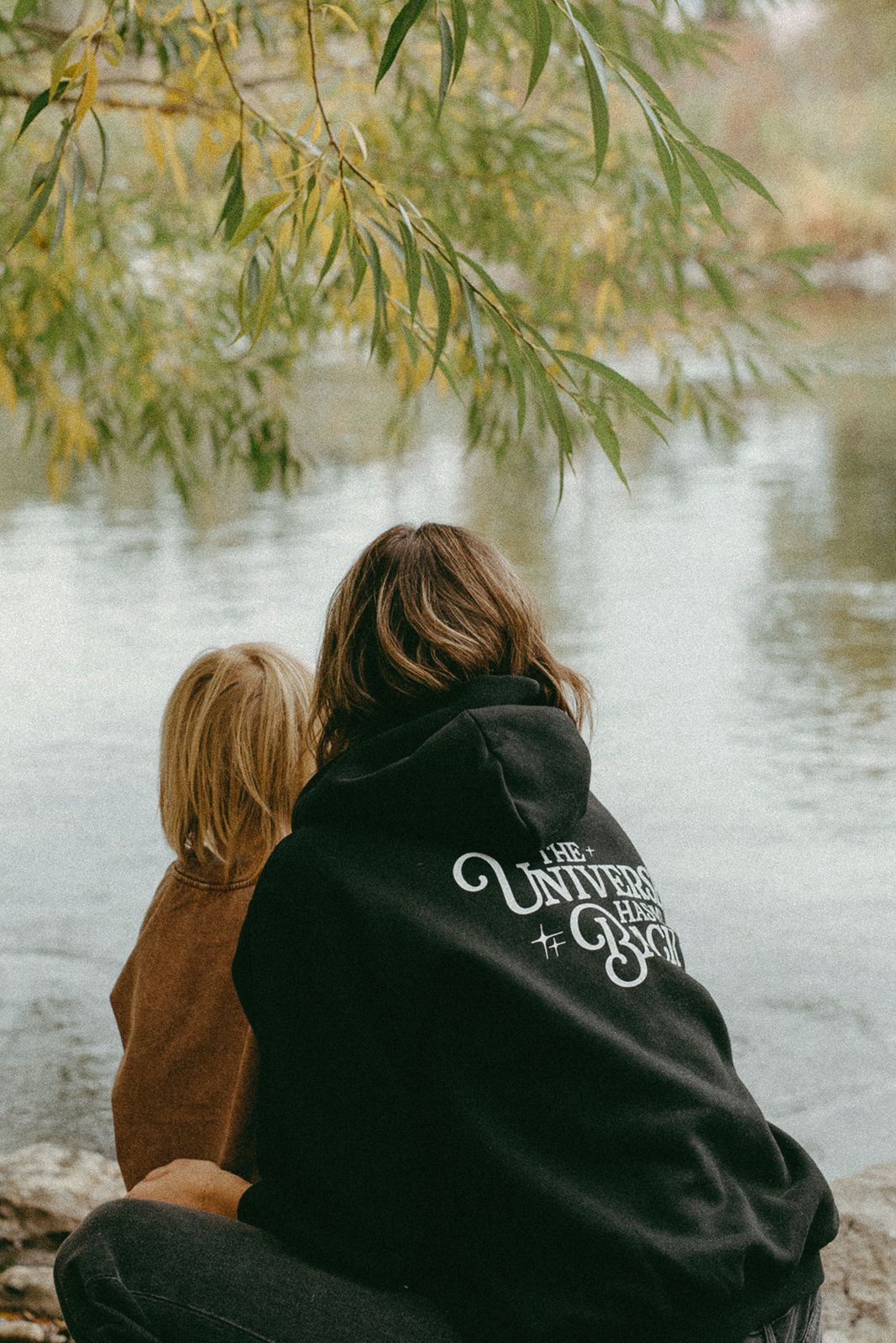 Woman and child sitting by a lake. They are wearing energy inspired and intentional apparel from the Dreamweavers collection. The woman is wearing a hoodie with the phrase "the universe has my back"