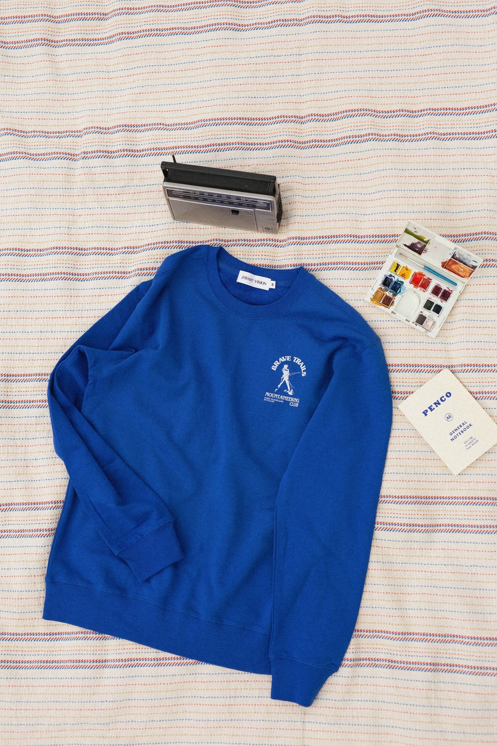 Front of blue sweatshirt with a "brave trails mountaineering" logo on a striped blanket background