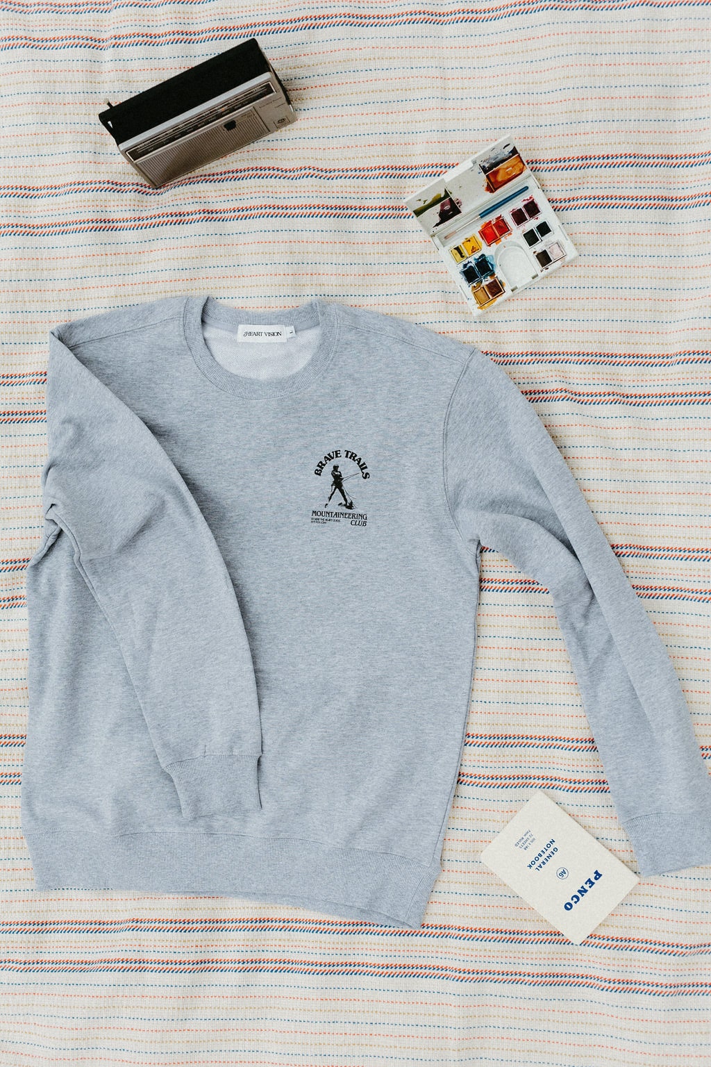 front of gray sweatshirt with brave trails mountaineering club logo on a striped blanket with paints and a radio