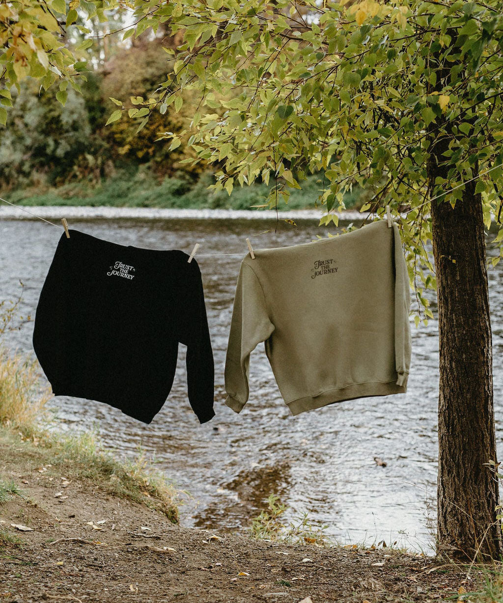 Two sweatshirts, one black and one khaki, hanging on a clothesline by a river with trees in the background.