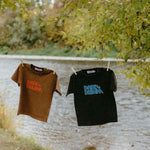 Two t-shirts, one brown and one black, with 'Dare to Dream' printed on them, hanging by a lake.