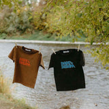 Two t-shirts, one brown and one black, with 'Dare to Dream' printed on them, hanging by a lake.