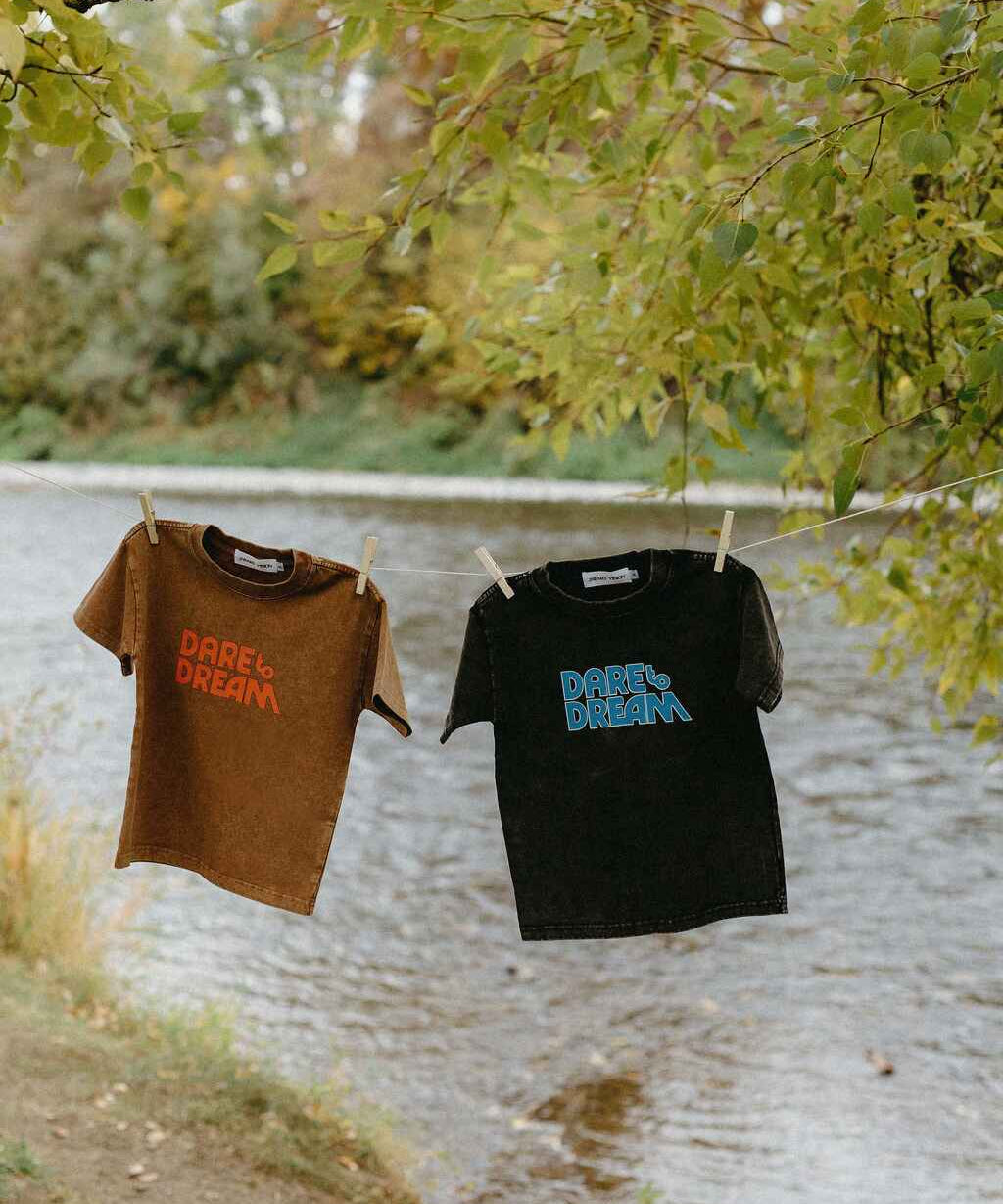 Two t-shirts, one brown and one black, with 'Dare to Dream' printed on them, hanging by a lake.