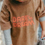 Child wearing a brown shirt with 'Dare to Dream' text, standing outdoors.