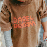 Child wearing a brown shirt with 'Dare to Dream' text, standing outdoors.