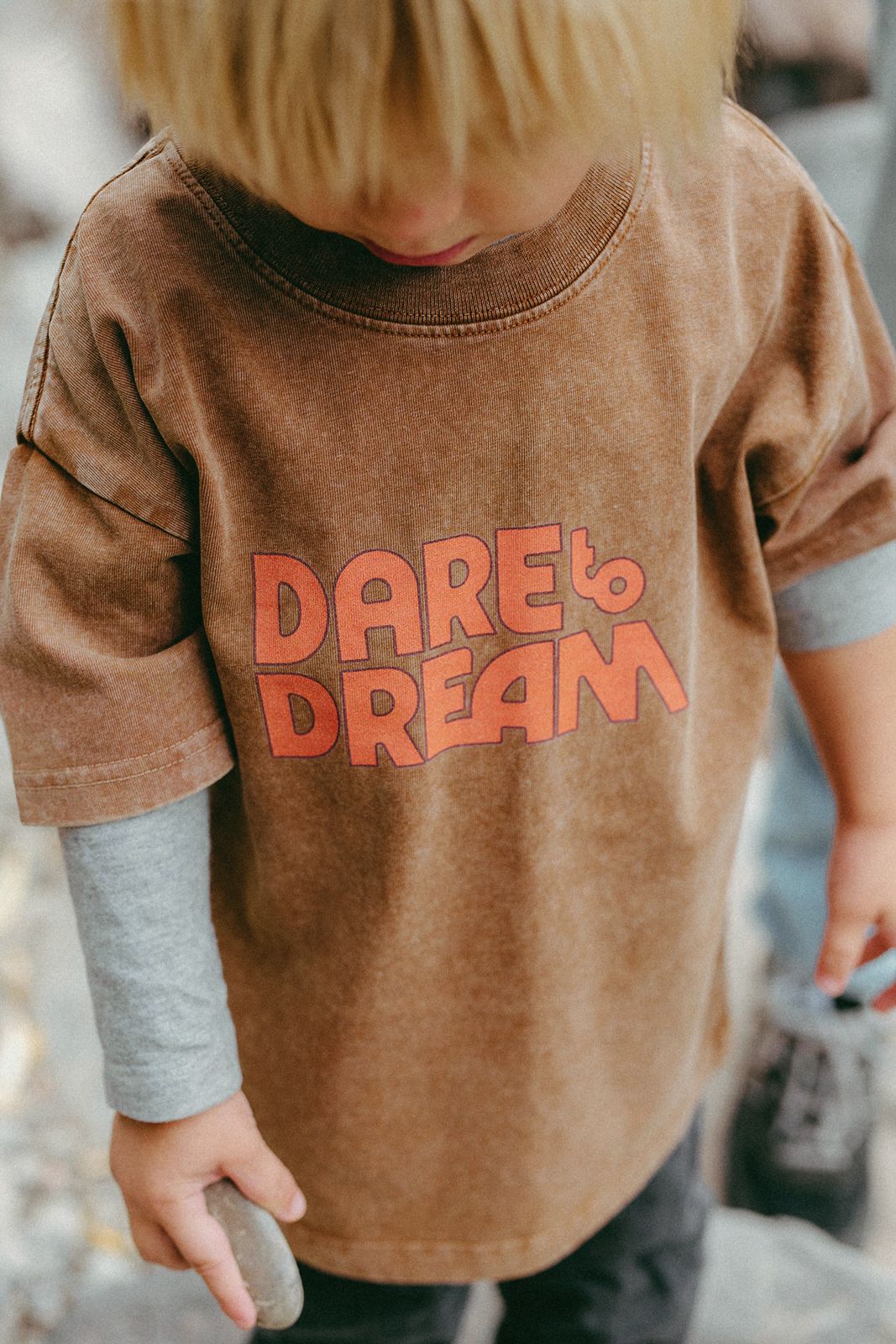 Child wearing a brown shirt with 'Dare to Dream' text, standing outdoors.