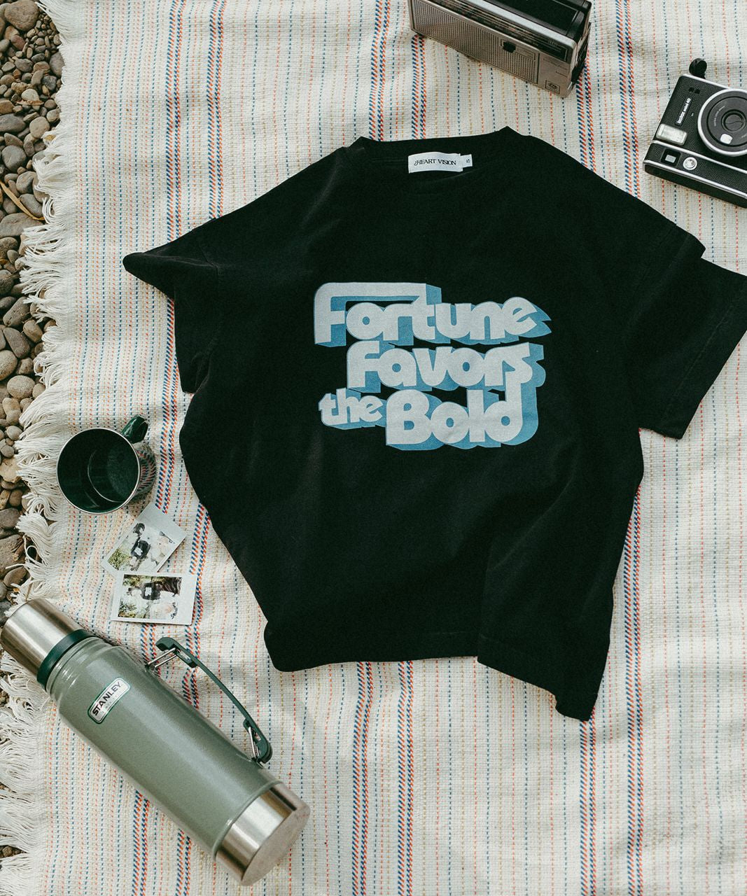 Black t-shirt with 'Fortune Favors the Bold' text on a striped blanket with a thermos and camera.