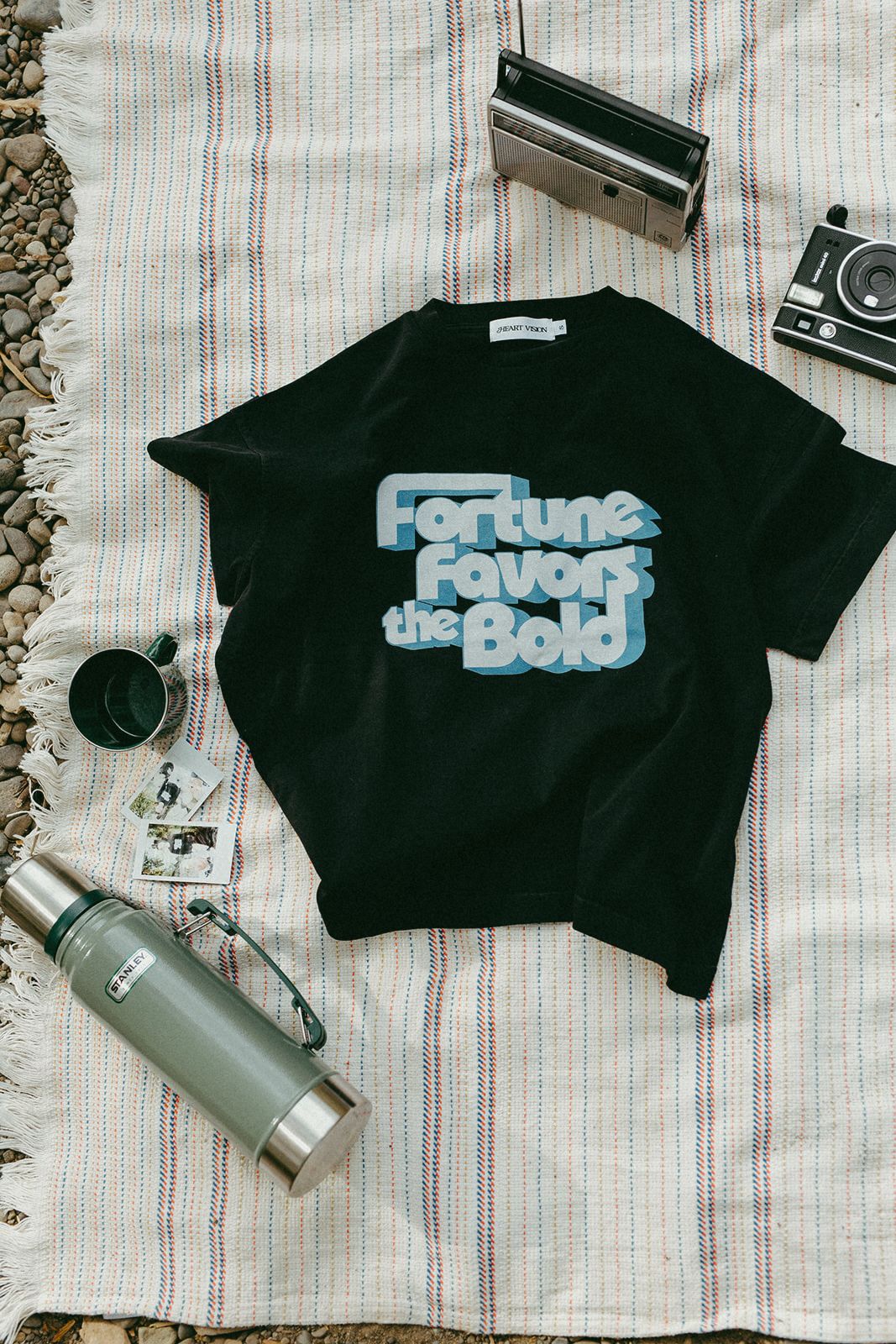Black t-shirt with 'Fortune Favors the Bold' text on a striped blanket with a thermos and camera.