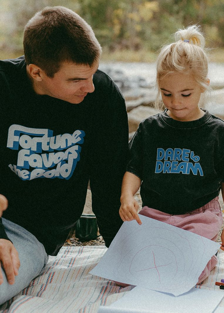 Man and young girl wearing black t-shirts with blue text, sitting outdoors on a blanket.