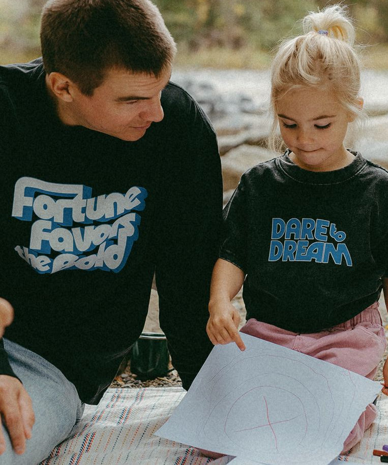 Man and young girl wearing black t-shirts with blue text, sitting outdoors on a blanket.
