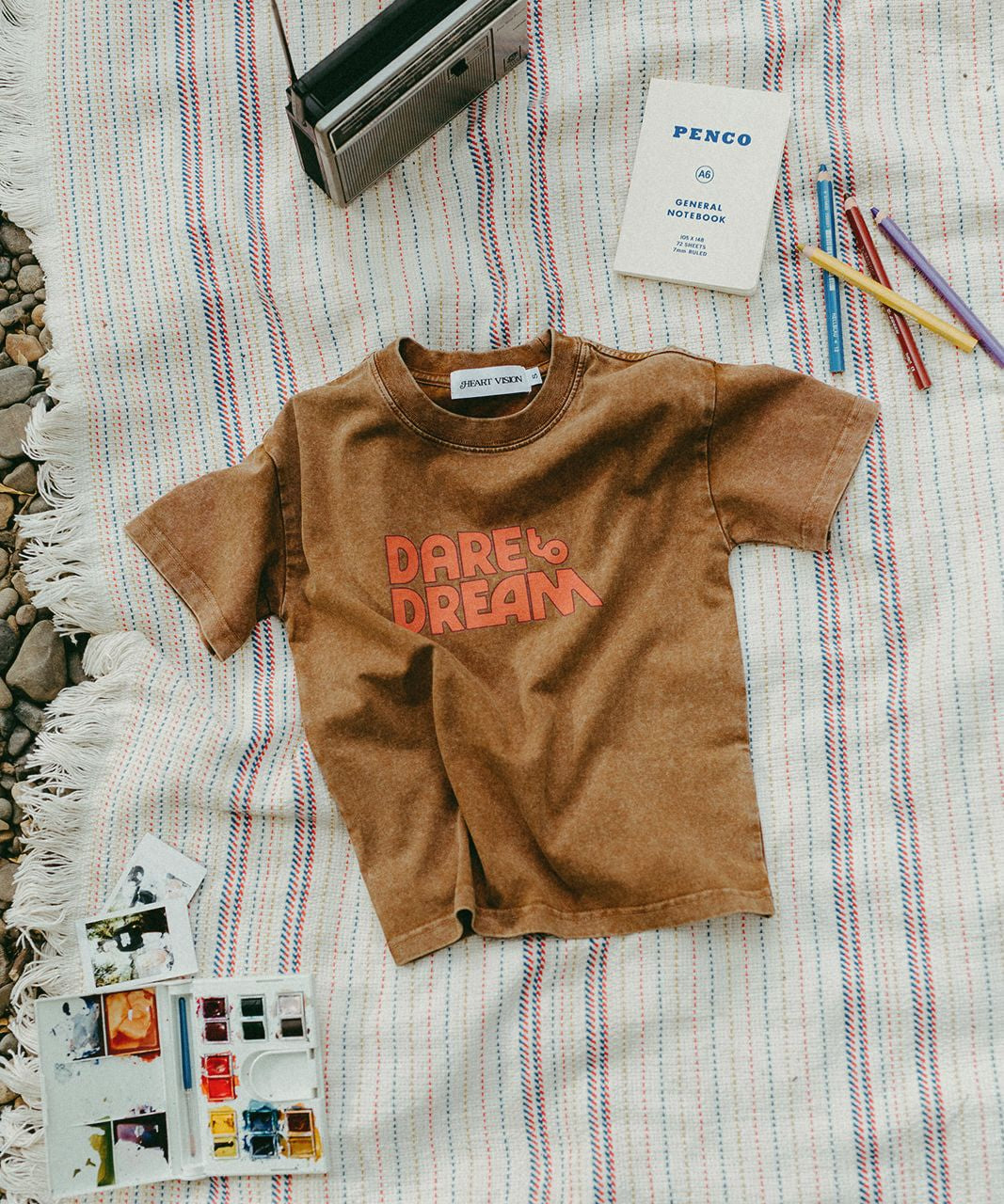 Brown t-shirt with 'Dare to Dream' text on a striped blanket with stationery items.