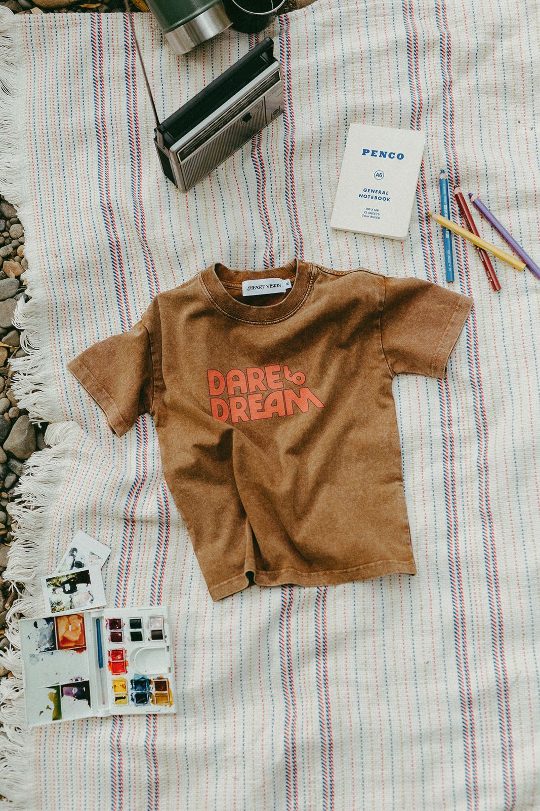 Brown t-shirt with 'Dare to Dream' text on a striped blanket with stationery items.