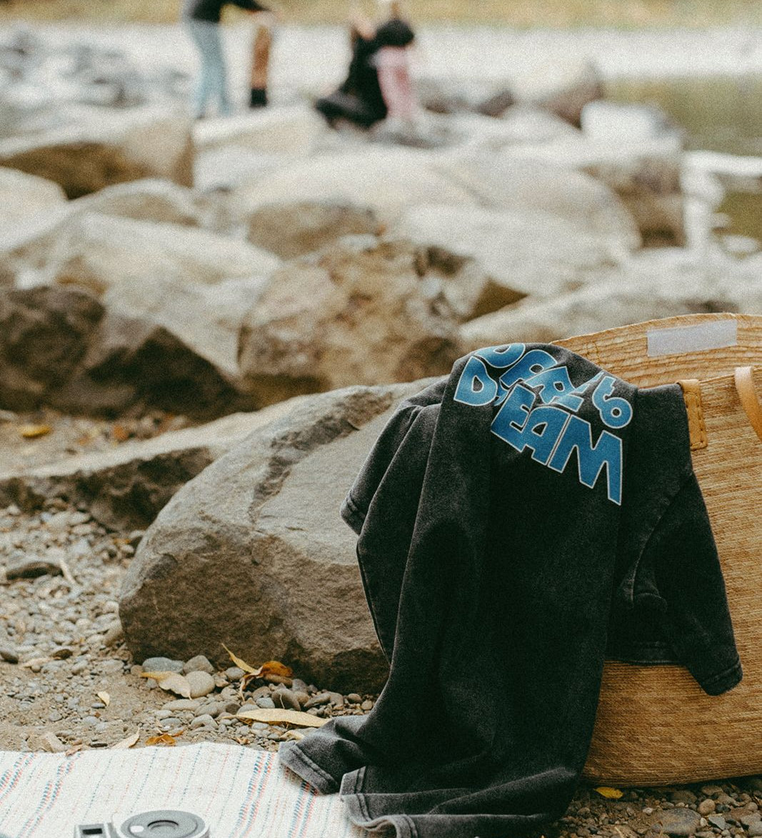Black t-shirt with 'dare to dream' text draped over a woven basket by a river with people in the background.