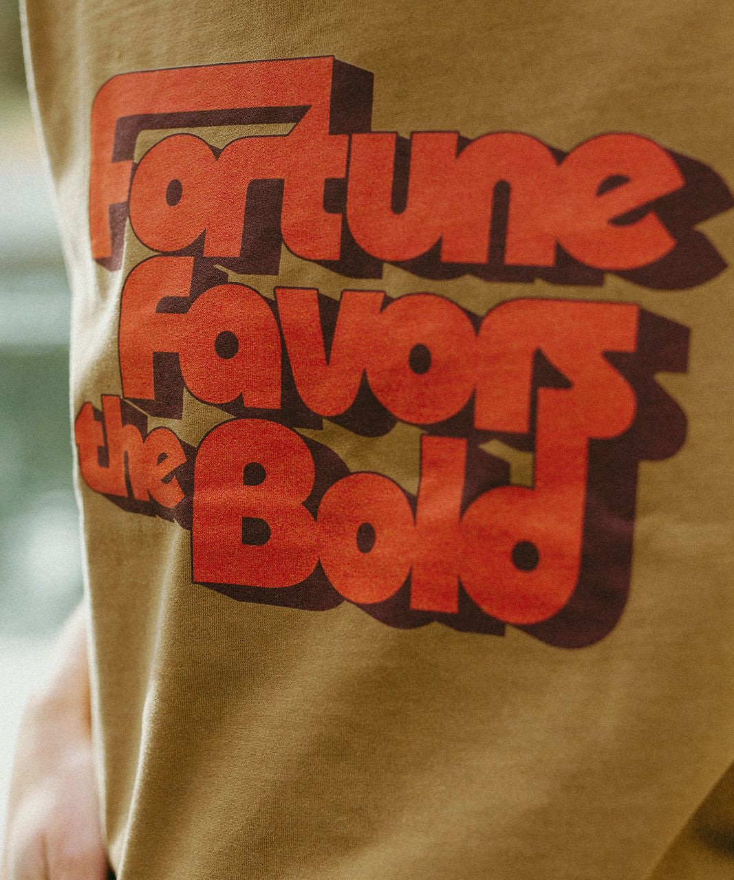 Brown t-shirt with red text 'Fortune Favours the Bold' on a blurred background