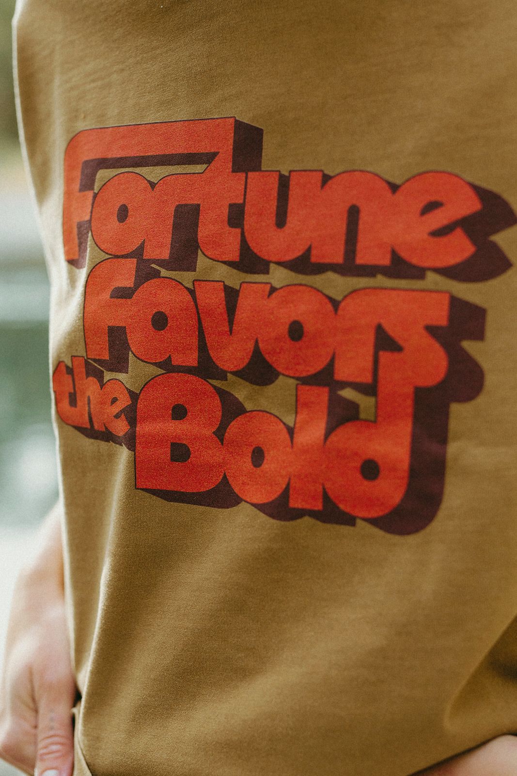 Brown t-shirt with red text 'Fortune Favours the Bold' on a blurred background