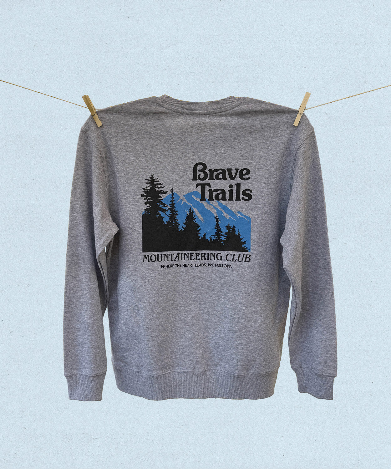 Back of gray sweatshirt with 'Brave Trails Mountaineering Club' design hanging on a clothesline on a light blue background