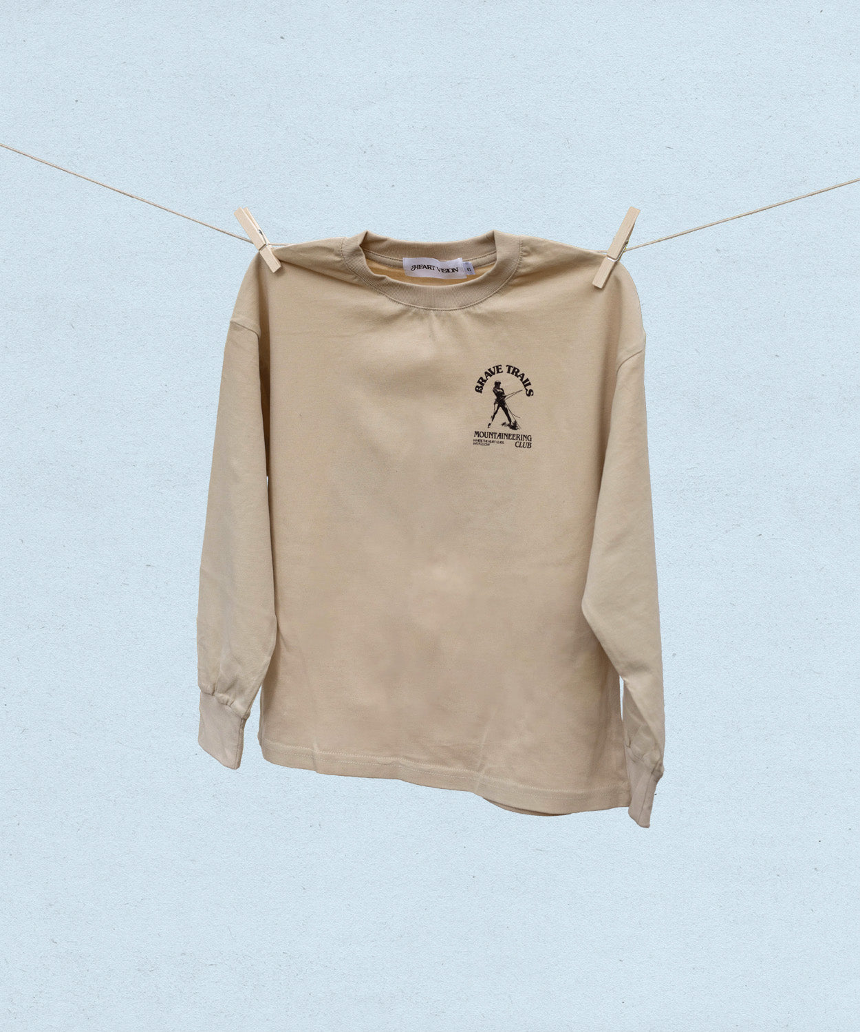 Beige long-sleeve t-shirt with a Brave Trails Mountaineering logo on a clothesline against a light blue background