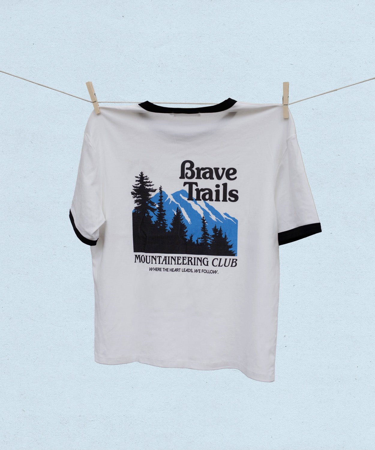 Back of white t-shirt with black and blue brave trails mountaineering graphic design hanging on a clothesline against a light blue background