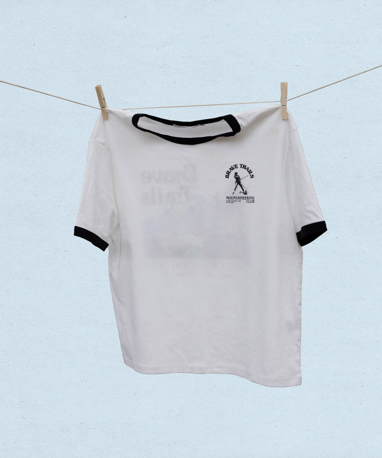 White t-shirt with black trim and a "brave trails mountaineering" logo hanging on a clothesline against a light blue background
