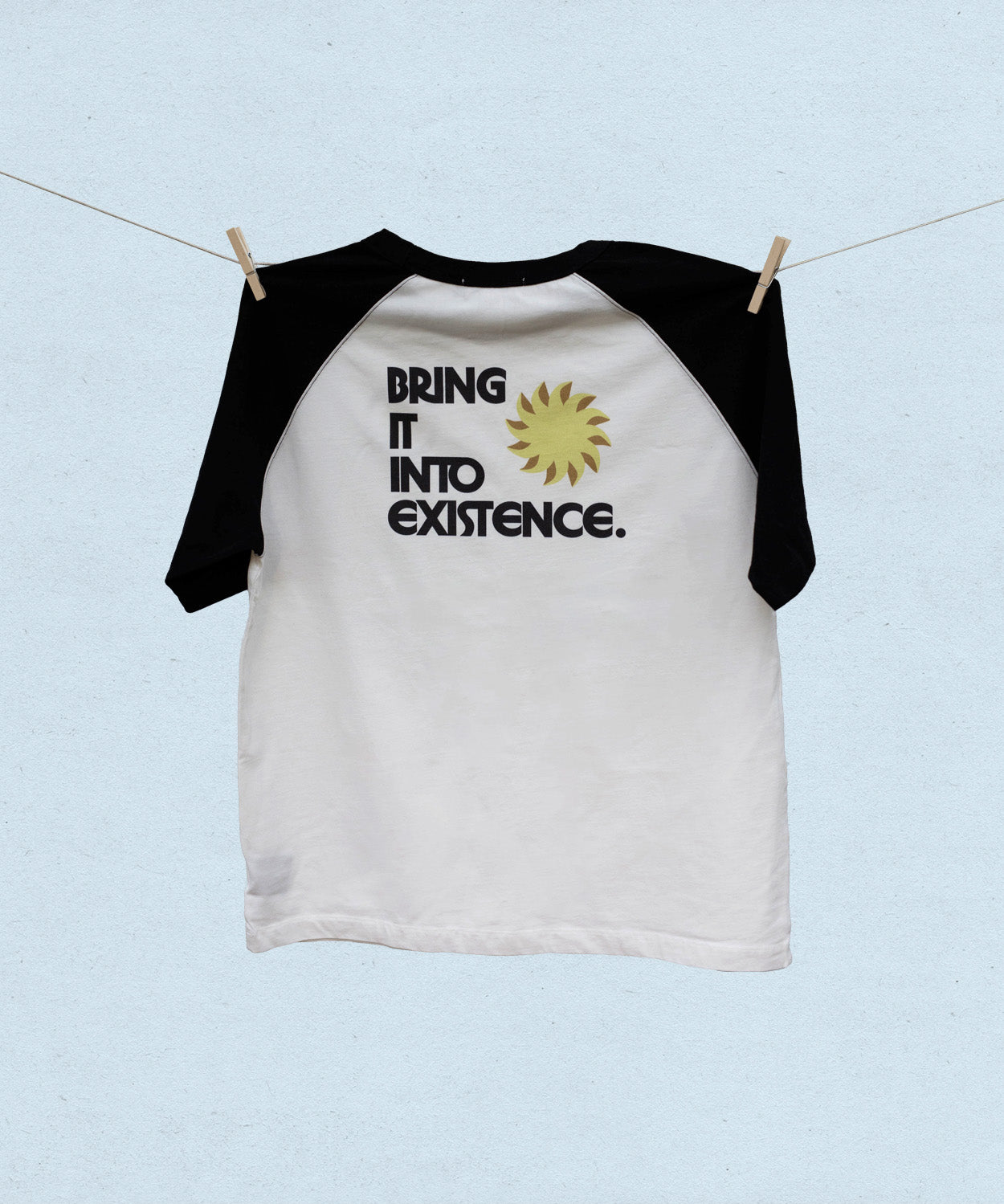Back of white baseball t-shirt with black short sleeves and text 'Bring it into existence' on a light blue background