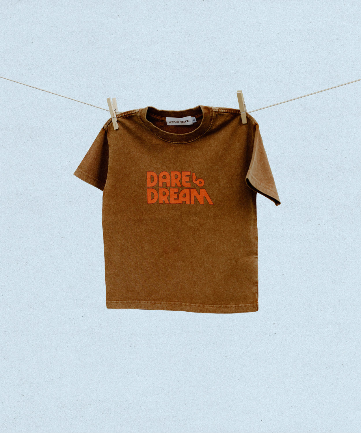 Brown t-shirt with red/orange 'Dare Dream' text on a clothesline against a light blue background