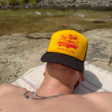 A person lying on a blanket by a river wearing a yellow trucker hat with a black brim and red graphic text reading 'Joy Ride state of mind'.