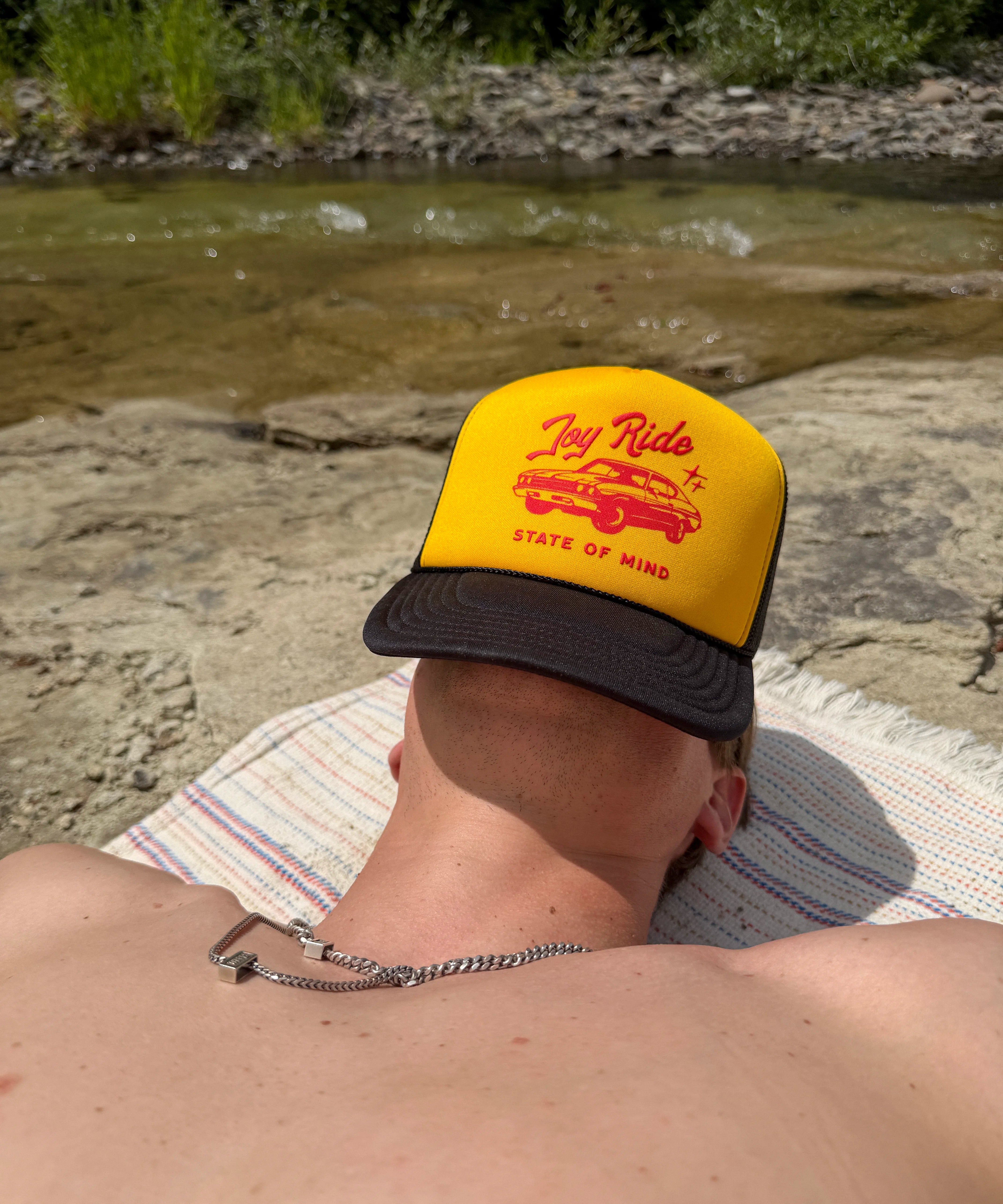 A person lying on a blanket by a river wearing a yellow trucker hat with a black brim and red graphic text reading 'Joy Ride state of mind'.