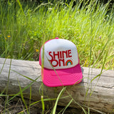 Shine On pink and white youth trucker hat with rainbow graphic resting on a log outdoors