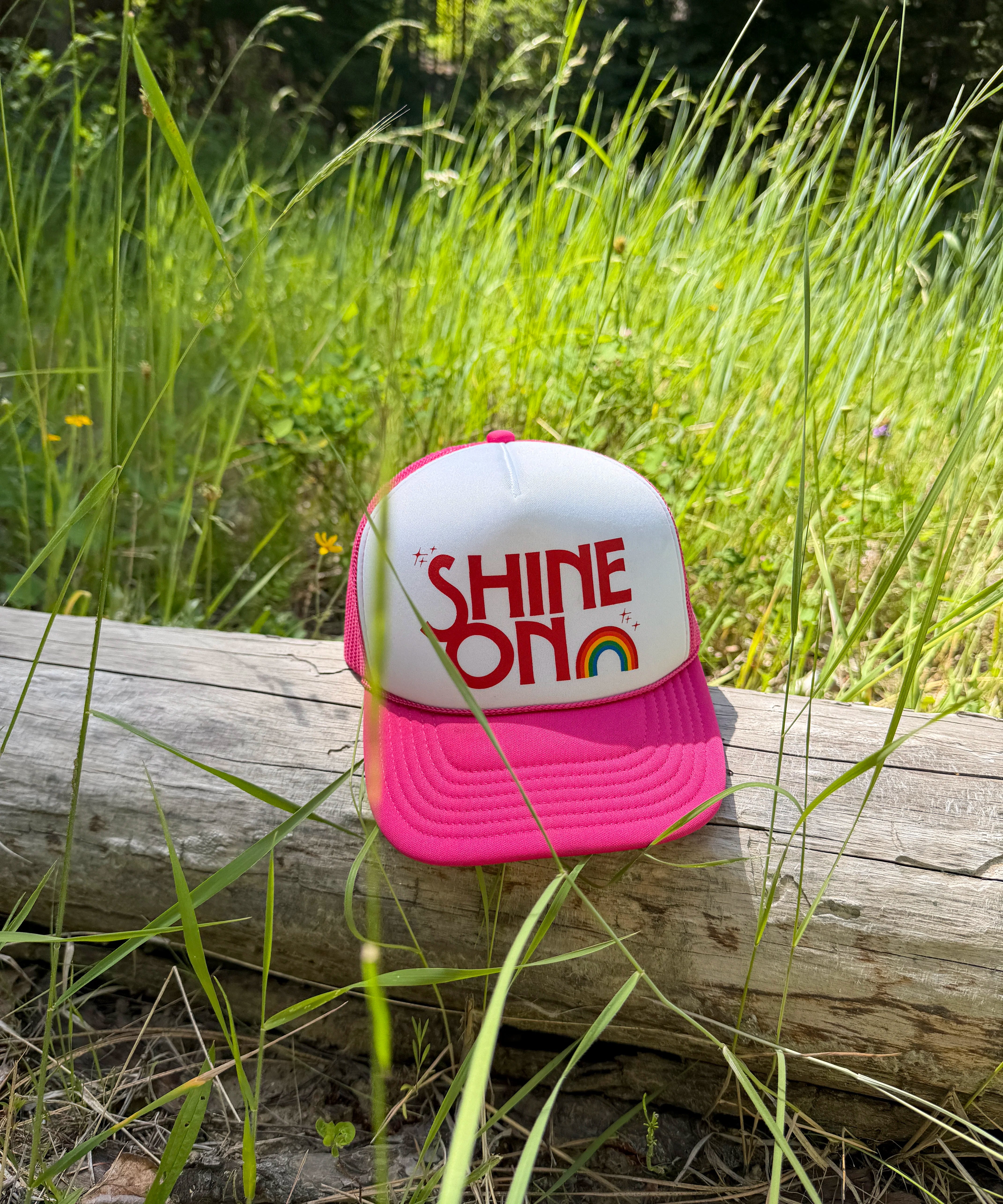 Shine On pink and white youth trucker hat with rainbow graphic resting on a log outdoors