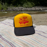 A yellow and black trucker hat with a red 'Joy Ride state of mind', resting on a striped blanket outdoors near a river and trees.